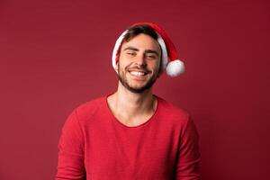 giovane bello caucasico tipo nel rosso maglione e Santa cappelli sta su rosso sfondo nel studio e e denti sorridente foto