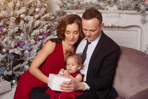 contento giovane famiglia con uno bambino Tenere Natale regalo. seduta su il divano vicino il Natale albero a casa foto