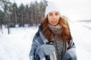 adorabile contento giovane bionda donna nel bianca a maglia cappello sciarpa avendo divertimento potabile caldo tè a partire dal thermos tazza nevoso inverno parco foresta nel natura foto