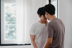 coppia gay asiatica in piedi e abbracciata vicino alla finestra di casa. i giovani uomini lgbtq asiatici che si baciano felici si rilassano riposano insieme trascorrono del tempo romantico nel soggiorno della casa moderna al mattino concetto. foto
