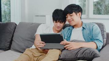 giovane coppia gay che utilizza tablet a casa. gli uomini lgbtq asiatici felici si rilassano e si divertono usando la tecnologia guardando film in internet insieme sdraiati sul divano nel concetto di soggiorno. foto