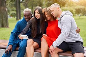 giovane persone di diverso nazionalità prendere selfie utilizzando smartphone, seduta su un' panchina nel il parco nel estate. atletico costruire. gli sport uniforme. foto