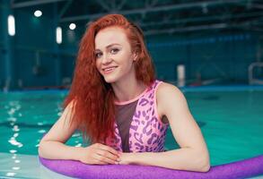 bellissimo giovane dai capelli rossi ragazza nel un' alla moda costume da bagno nel il stile di il anni 80 sta con il tagliatelle nel sua mani nel il interno nuoto piscina foto