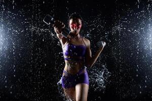 bellissimo giovane ragazza nel viola abbigliamento sportivo pose con manubri nel acqua studio. gocce di acqua diffusione di sua fitness corpo. il Perfetto figura su il sfondo di acqua spruzzi foto