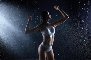 bellissimo giovane ragazza nel abbigliamento sportivo pose nel acqua studio. gocce di acqua diffusione di sua fitness corpo. il Perfetto figura su il sfondo di acqua spruzzi foto