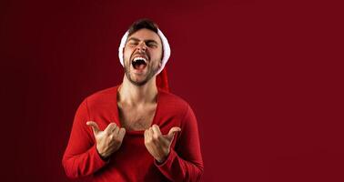 giovane bello caucasico tipo nel rosso maglione e Santa cappelli sta su rosso sfondo nel studio Spettacoli Come caldo lui è foto