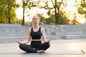 yoga e meditazione nel moderno città caucasico donna rilassare loto posizione seduta yoga stuoia all'aperto estate parco su calcestruzzo pavimento foto