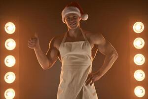 caldo Natale dolce. muscolare uomo indossare grembiule e Santa cappello mostrando pollice su gesto e sorridente a telecamera, lampade illuminazione su sfondo foto