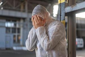 disperazione assistenza sanitaria lavoratore nel bianca covid protettivo tuta da lavoro. stanco maschio caucasico medico afferra il suo testa rannicchiarsi nel depressione durante coronavirus pandemia foto