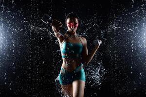 bellissimo giovane ragazza nel abbigliamento sportivo pose con manubri nel acqua studio. gocce di acqua diffusione di sua fitness corpo. il Perfetto figura su il sfondo di acqua spruzzi foto