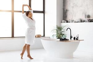 bellissimo nero femmina nel bianca accappatoio rilassante e allungamento nel moderno bagno foto