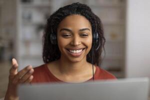 sorridente giovane nero donna con cuffia comunicare di video chiamata su il computer portatile foto