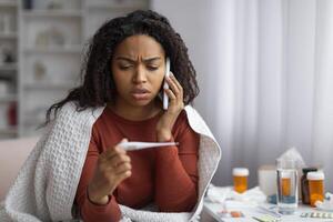 in linea consultazione. malato nero donna Tenere termometro e chiamata medico su cellulare foto