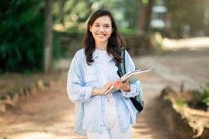 contento giovane donna con un' affascinante Sorridi Tenere i Quaderni mentre con fiducia a piedi nel un' parco foto