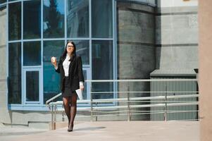 ritratto di riuscito attività commerciale donna andando per opera con caffè a piedi vicino ufficio edificio su città strada. foto