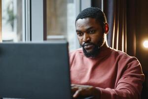 ritratto di contento africano uomo d'affari utilizzando Telefono mentre Lavorando su il computer portatile nel ristorante. foto