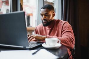 profilo ritratto di giovane africano in linea negozio gestore, parlando su mobile Telefono per cliente, consulenza lui su consegna termini, seduta a bar e utilizzando il computer portatile computer per opera, avendo caffè e torta. foto