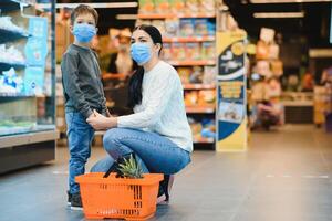 shopping con ragazzo durante virus scoppio. madre e bambino indossare chirurgico viso maschera acquisto frutta nel supermercato. mamma e poco ragazzo acquistare fresco verdura nel drogheria negozio. famiglia nel negozio foto