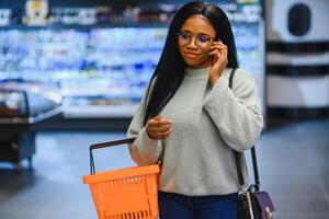 africano americano donna con shopping carrello carrello nel il supermercato memorizzare parlare su mobile Telefono foto