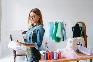 sorridente moda progettista guardare a telecamera a posto di lavoro, sarta, ricamatrice o sarto negozio proprietario seduta a scrivania con colore campioni pantone e ricamo design schizzi su il parete. foto