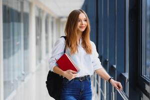 allegro brunetta alunno ragazza con nero zaino detiene libri nel moderno costruzione. femmina alunno in piedi con libri nel Università corridoio foto