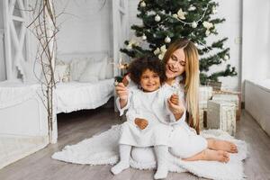 allegro Natale e contento vacanze allegro mamma e sua carino figlia a Natale albero. genitore e poco bambino avendo divertimento vicino Natale albero al chiuso. amorevole famiglia con regali nel camera. foto