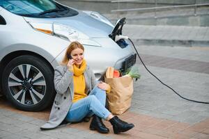 utilizzando smartphone mentre in attesa. donna su il elettrico macchine caricare stazione a giorno. marca nuovo veicolo foto