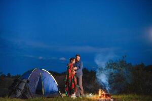 notte campeggio nel il montagne. contento coppia i viaggiatori seduta insieme accanto fuoco di bivacco e raggiante turista tenda. su sfondo grande masso, foresta e notte cielo. foto