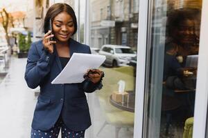 accigliato ha riguardato manager A proposito di su cellulare fuori. giovane africano americano attività commerciale donna in piedi vicino all'aperto bicchiere parete. mobile Telefono parlare concetto foto