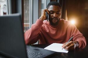 profilo ritratto di giovane africano in linea negozio gestore, parlando su mobile Telefono per cliente, consulenza lui su consegna termini, seduta a bar e utilizzando il computer portatile computer per opera, avendo caffè e torta. foto