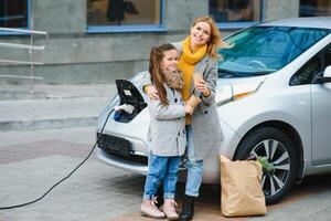 madre con figlia ricarica elettro auto a il elettrico gas stazione e parlare su mobile Telefono foto