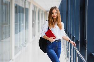 positività bellissimo ragazza sorridente a telecamera, in piedi su corridoio con Appunti come zaino, andando per lezione. contento brunetta femmina alunno studiando nel lusso Università. foto