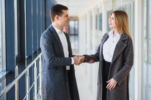 attività commerciale uomini e donne tremante mani con un' Sorridi su il sfondo di il grande panoramico finestre nel un' moderno attività commerciale centro. Modelli vestito nel un' buio attività commerciale abiti. foto