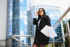 giovane ragazza ufficio lavoratore nel occhiali, buio giacca e blu camicia soggiorni su città strada vicino ufficio edificio e detiene sua documenti. foto