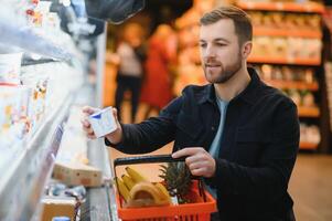 cliente nel supermercato. uomo fare drogheria shopping in piedi con carrello la scelta cibo Prodotto al chiuso. tipo acquisto drogheria nel cibo negozio. selettivo messa a fuoco, copia spazio. foto