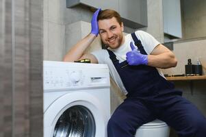 il giovane bello riparatore nel lavoratore completo da uomo con il professionale utensili scatola è fissaggio il lavaggio macchina nel il bagno foto