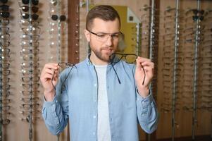 nel ottica negozio. ritratto di maschio cliente Tenere e indossare diverso spettacoli, la scelta e provando su nuovo bicchieri a ottico negozio. uomo raccolta telaio per visione correzione, avvicinamento foto