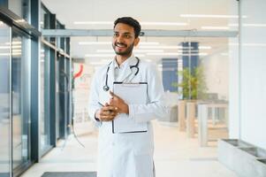 ritratto di maschio indiano medico indossare bianca cappotto avendo Aperto porta su clinica corridoio come sfondo foto