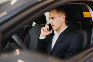 giovane uomo d'affari si siede nel lusso auto e parla su Telefono. lui sembra dritto inoltrare. tipo unità macchina. lui detiene uno mano su timone ruota. esso è soleggiato fuori. foto