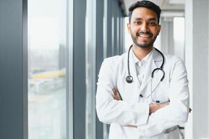 ritratto di contento amichevole maschio indiano latino medico medico lavoratore indossare bianca cappotto con stetoscopio in giro collo in piedi nel moderno privato clinica guardare a telecamera. medico assistenza sanitaria concetto foto