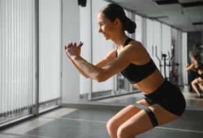 motivazione per ragazze. moderno giovane donna nel sport capi di abbigliamento accovacciato utilizzando resistenza gruppo musicale mentre esercizio nel il palestra. foto