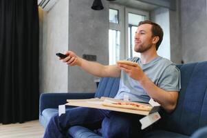 riposo dopo difficile opera. un' uomo è seduta su il divano a casa, mangiare Pizza e Guardando tv. foto