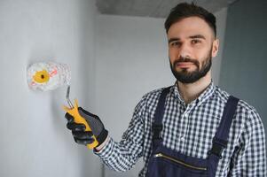 giovane sorridente professionale lavoratore nel blu uniforme in piedi con dipingere rullo nel nuovo appartamento per riparazione al di sopra di grigio muri sfondo, copia spazio. foto