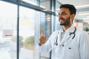 ritratto di maschio indiano medico indossare bianca cappotto avendo Aperto porta su clinica corridoio come sfondo foto