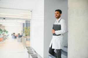 ritratto di maschio indiano medico indossare bianca cappotto avendo Aperto porta su clinica corridoio come sfondo foto