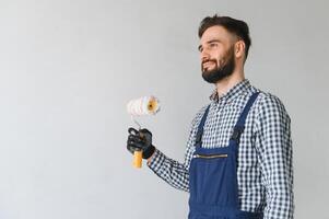 giovane sorridente professionale lavoratore nel blu uniforme in piedi con dipingere rullo nel nuovo appartamento per riparazione al di sopra di grigio muri sfondo, copia spazio. foto