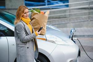 elegante donna dopo acquisto prodotti con un' shopping Borsa è in piedi vicino il ricarica elettrico macchina. foto