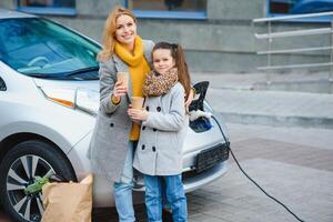 madre con figlia ricarica elettro auto a il elettrico gas stazione e parlare su mobile Telefono foto