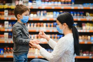 madre si applica disinfettante per pulizia figli maschi mani nel pubblico affollato posto. shopping centro commerciale foto