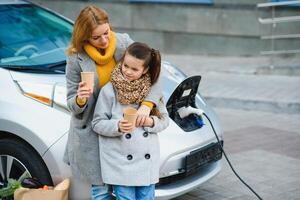 madre con figlia ricarica elettro auto a il elettrico gas stazione e parlare su mobile Telefono foto